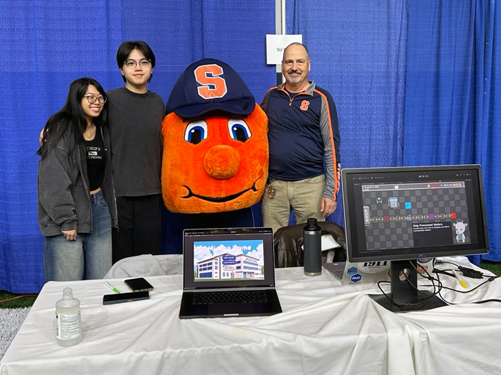 A woman, an orange mascot, and a man standing behind a table with computers on top.